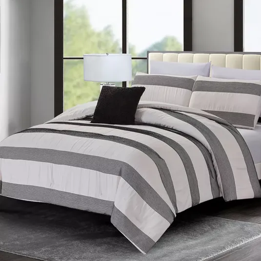 A bedroom features a bed with a striped comforter set in black and white, complemented by matching pillows and a black accent pillow. A table lamp sits beside the bed on a side table, all set against large windows and a plush gray rug.