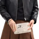 A person wearing a black leather jacket and brown plaid skirt holds a clutch styled like a vintage Coach advertisement, featuring text and graphics.
