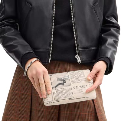 A person wearing a black leather jacket and brown plaid skirt holds a clutch styled like a vintage Coach advertisement, featuring text and graphics.