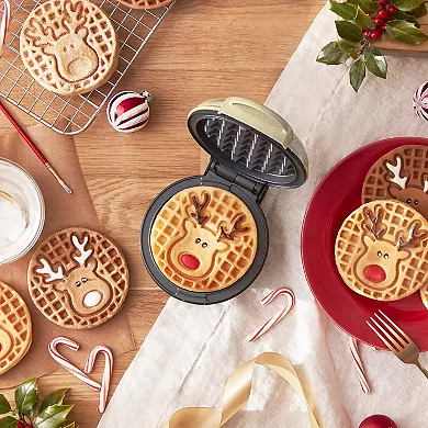 A mini waffle maker with a reindeer design imprint is open, displaying a freshly cooked waffle, surrounded by similarly designed waffles on a festive table setting.