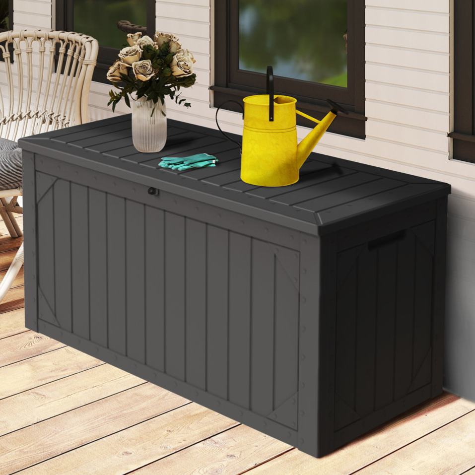 A dark outdoor storage bench with a lattice pattern is on a wooden deck. It holds a yellow watering can, gardening gloves, and a vase with flowers. A wicker chair and green plants are nearby.