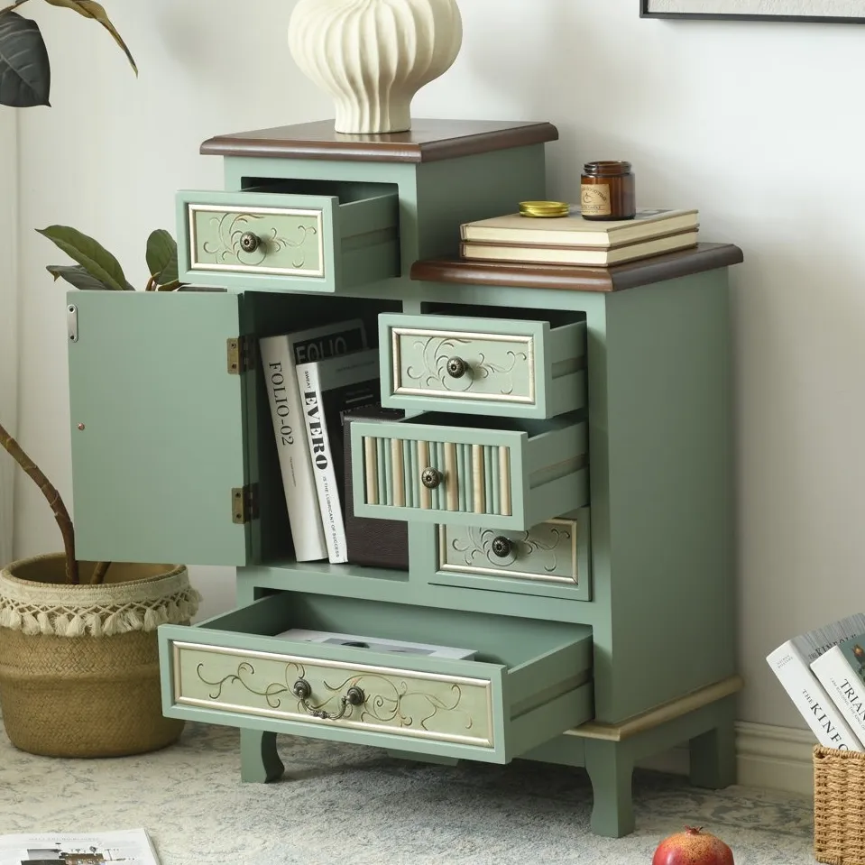A green vintage-style cabinet with several open drawers and a small cupboard, adorned with decorative knobs. It sits on a patterned rug, surrounded by books, a candle, and a vase on top.