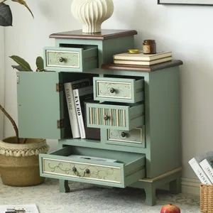 A green vintage-style cabinet with several open drawers and a small cupboard, adorned with decorative knobs. It sits on a patterned rug, surrounded by books, a candle, and a vase on top.