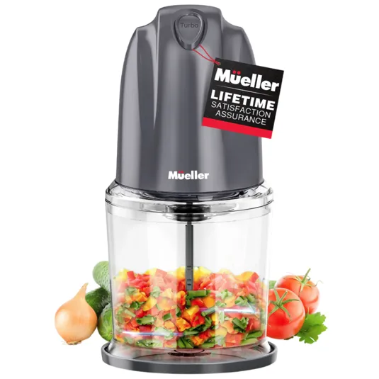 Mueller electric food chopper with turbo button, containing chopped vegetables. Includes \“Lifetime Satisfaction Assurance\“ tag and surrounded by fresh produce like tomatoes, onion, and cucumbers.