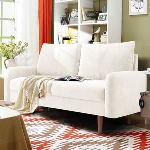A cream-colored 72\“ velvet plush sofa with wooden legs features two large cushions, a folded throw blanket, an open book, and a bold patterned rug. Cozy and simple, it's placed in a bright living room with a window and a bookshelf nearby.