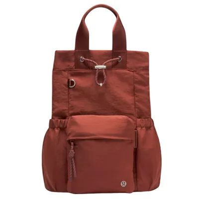 A rust-colored tote backpack with short handles, a front pocket, drawstring closure, and side pockets. It features a small circular logo at the front.
