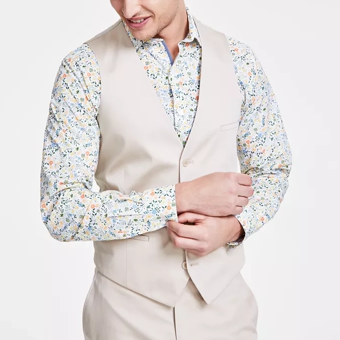 A slim-fit suit vest in a solid beige color featuring a V-neckline, button-front closure, and welt pockets. The model is wearing a floral shirt underneath.
