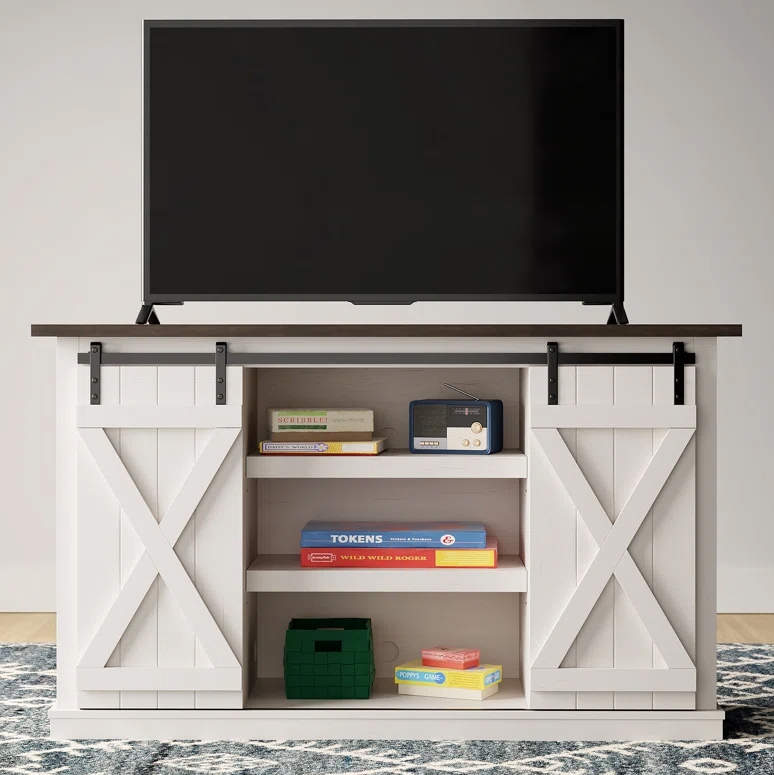 White TV stand with sliding barn doors, dark wood top, and three shelves holding books, a small radio, a green basket, and board games.