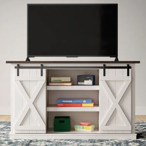 White TV stand with sliding barn doors, dark wood top, and three shelves holding books, a small radio, a green basket, and board games.