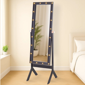 A black standing mirror with built-in lights on the frame is placed in a bedroom setting, near a bed and plants.