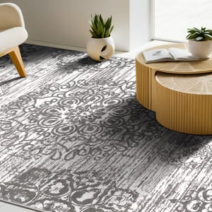 A patterned rug with intricate designs lies on the floor, accompanied by a wooden round coffee table with a book on top. There are potted plants nearby, and part of a white armchair is visible.