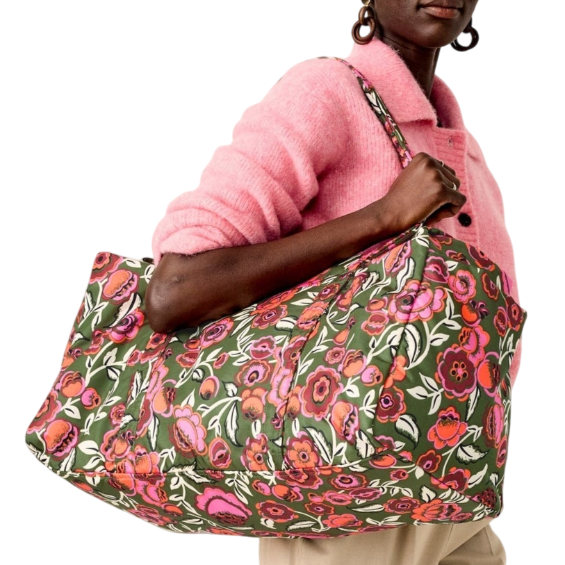 A person is holding a large tote bag featuring a colorful floral pattern with pink and red flowers on a green background, while wearing a pink sweater and beige pants.