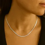 A woman's neck adorned with a thin, sleek silver necklace, possibly a flat snake chain, over a dark garment.