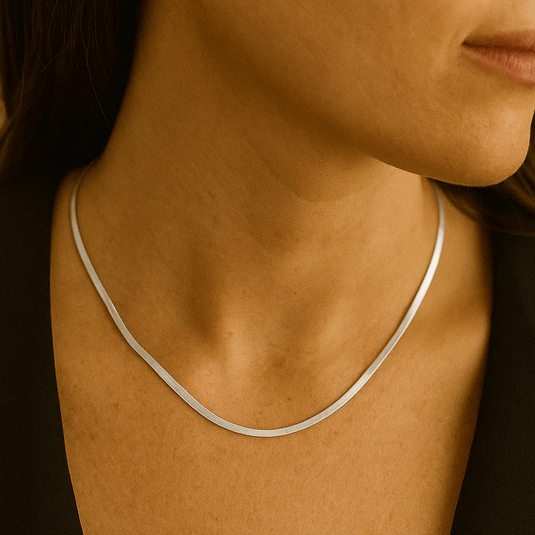 A woman's neck adorned with a thin, sleek silver necklace, possibly a flat snake chain, over a dark garment.