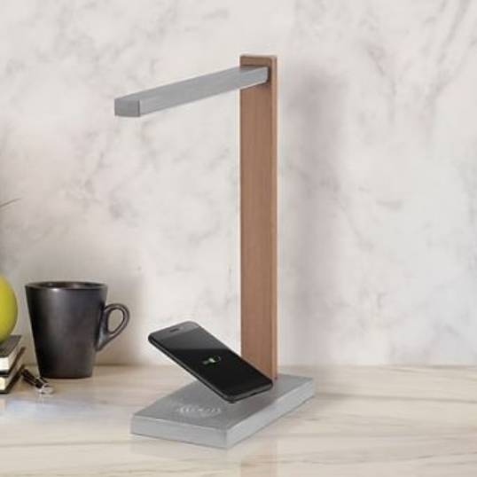 A modern desk lamp with a wooden stand features a built-in wireless charging pad, which is currently charging a smartphone. Nearby are a black mug and a stack of books on a marble-like surface.