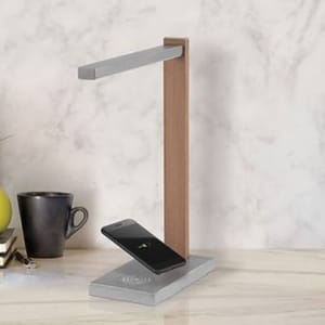A modern desk lamp with a wooden stand features a built-in wireless charging pad, which is currently charging a smartphone. Nearby are a black mug and a stack of books on a marble-like surface.