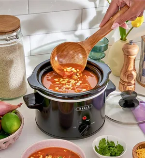 A slow cooker with tomato and corn soup is on a kitchen counter, accompanied by a wooden ladle. Nearby are a glass jar of rice, limes, shallot, fresh cilantro, and a pepper grinder.