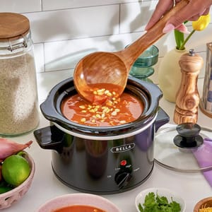 A slow cooker with tomato and corn soup is on a kitchen counter, accompanied by a wooden ladle. Nearby are a glass jar of rice, limes, shallot, fresh cilantro, and a pepper grinder.