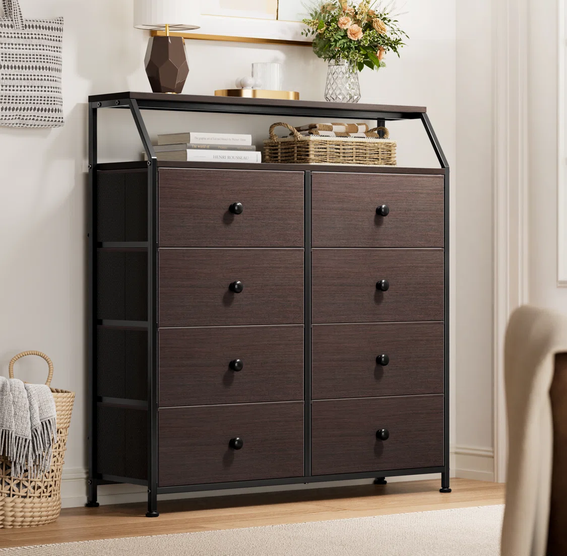 A dark brown dresser with eight drawers featuring black knobs stands on a wooden floor. It has a metal frame, and is topped with a shelf displaying a lamp, books, a basket, and flowers in a vase.