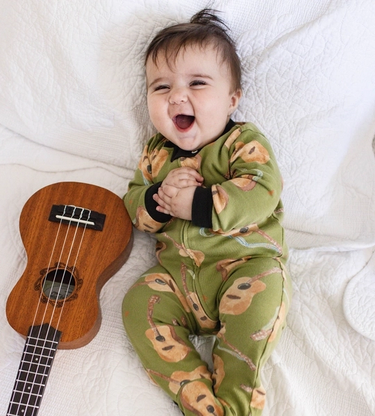 A baby wearing green pajamas with bear designs is lying on a white blanket, smiling, with a ukulele beside them.