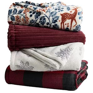 A stack of four cozy blankets featuring various designs: a floral-and-deer pattern, a solid burgundy, a snowflake print, and a red-and-black checkered pattern.