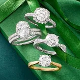 Four diamond engagement rings are displayed on a green gift box wrapped with ribbon: three with white metal bands and one with a gold band.
