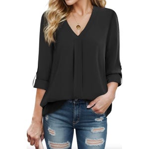 A woman is wearing a black V-neck blouse with 3/4 sleeves, paired with distressed blue jeans. She's holding a light-colored clutch and accessorized with a round pendant necklace.