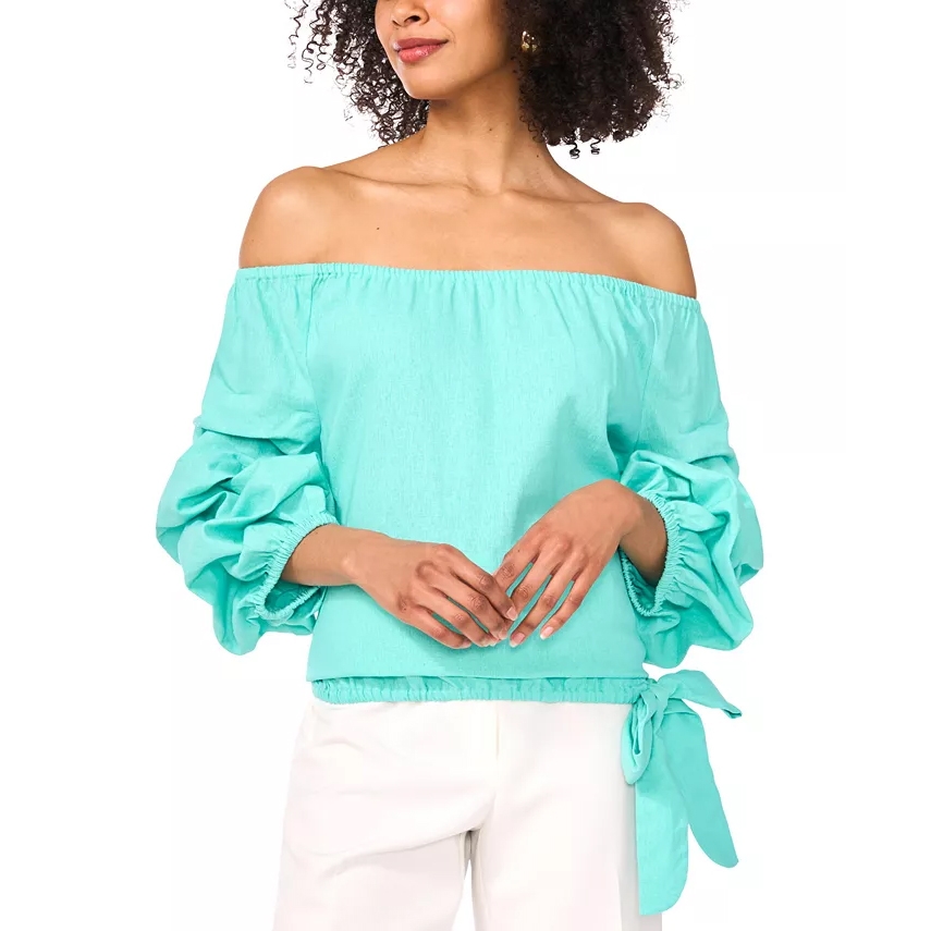 A woman is wearing a turquoise off-the-shoulder blouse with voluminous sleeves and a side tie, paired with white pants.