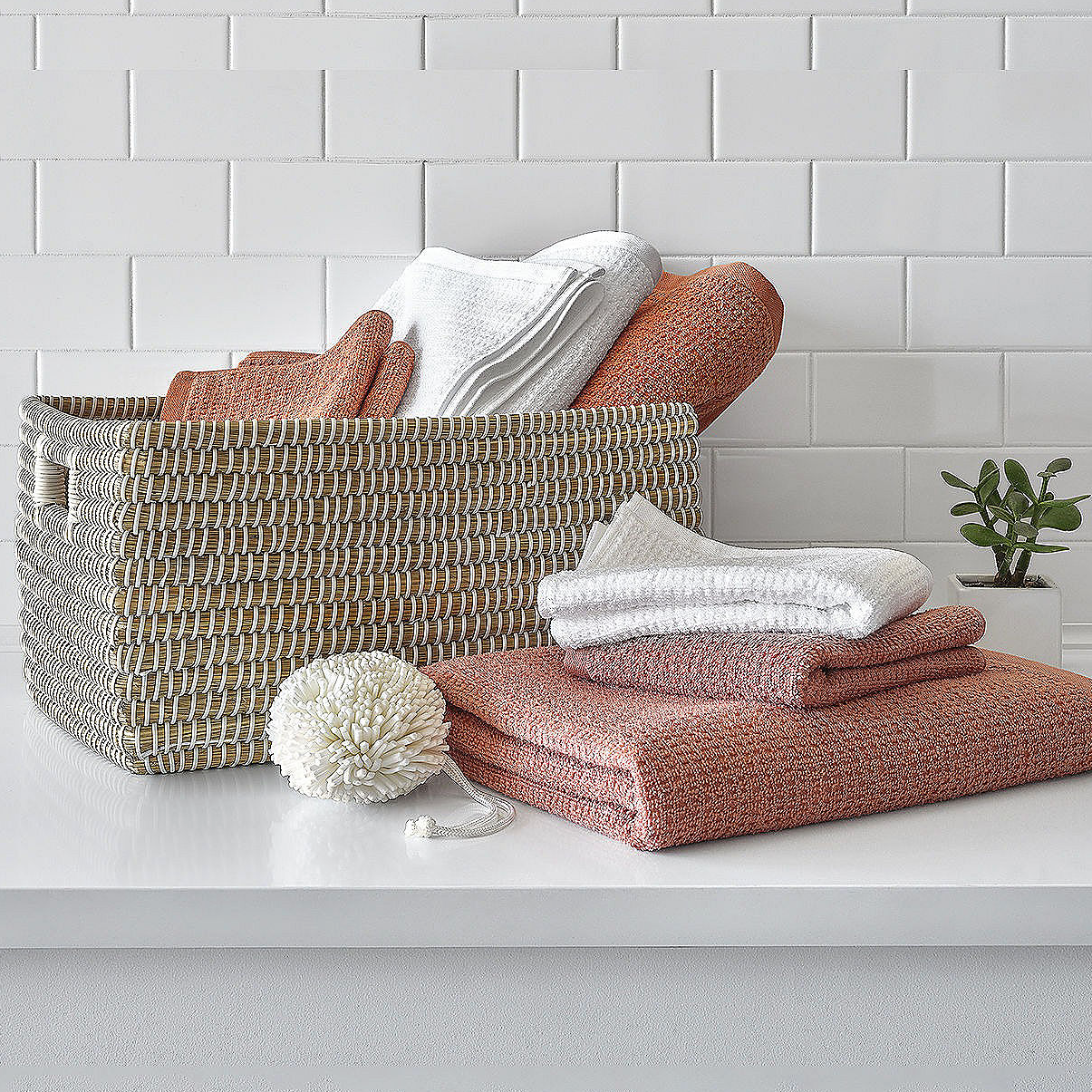 Woven basket with textured bath towels in shades of coral and white and a decorative white pom-pom on a white surface against a brick wall.