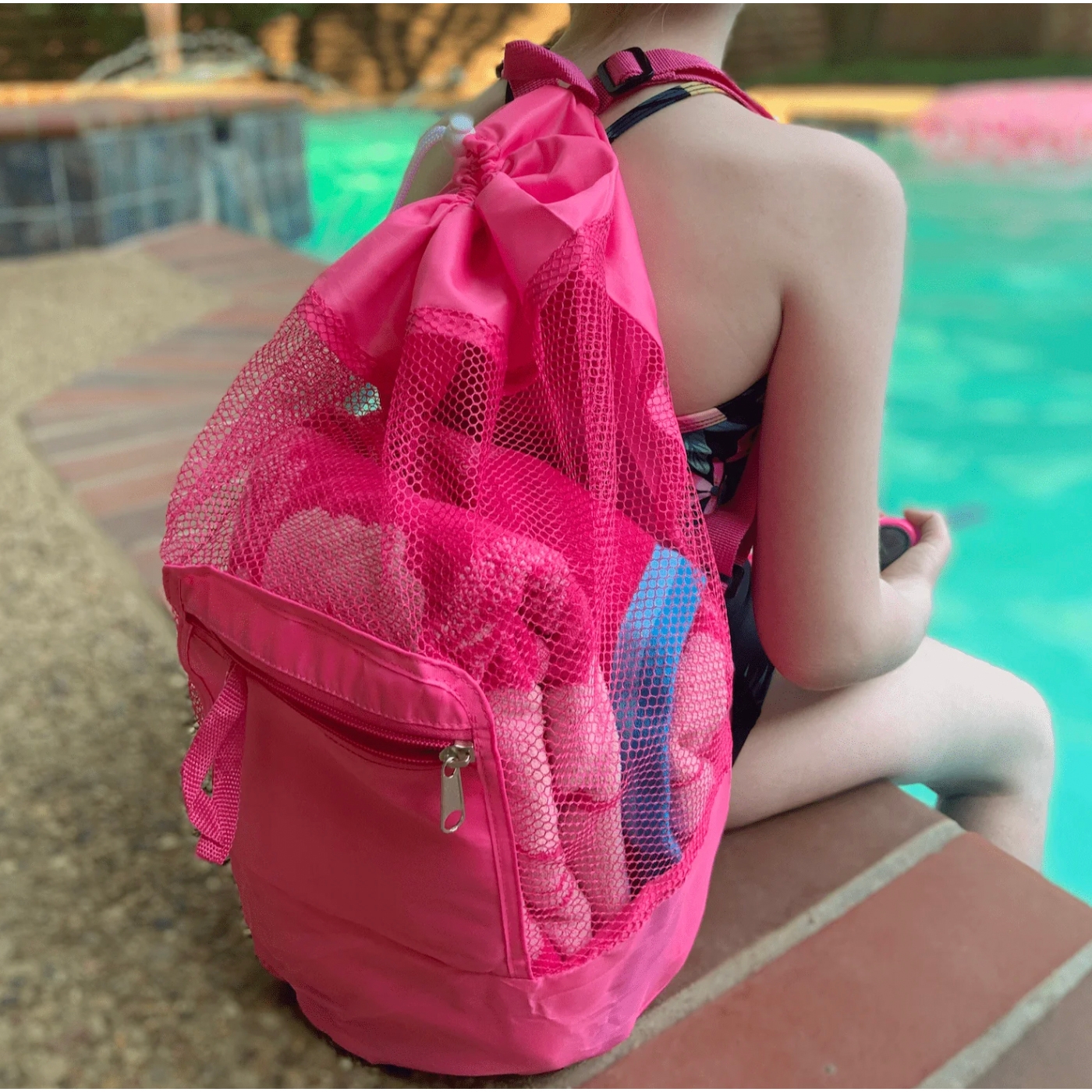 A pink mesh backpack with a drawstring closure and zipper pocket, containing rolled-up towels, is worn by a person sitting by a pool.