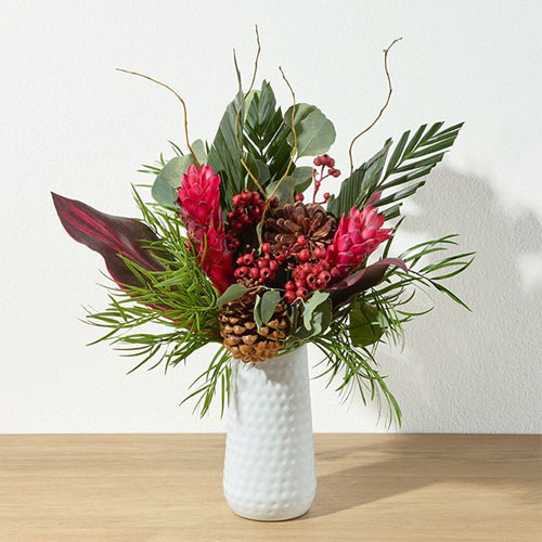 A vibrant floral arrangement in a white textured vase features pink flowers, pinecones, and assorted greenery with a natural, organic look.