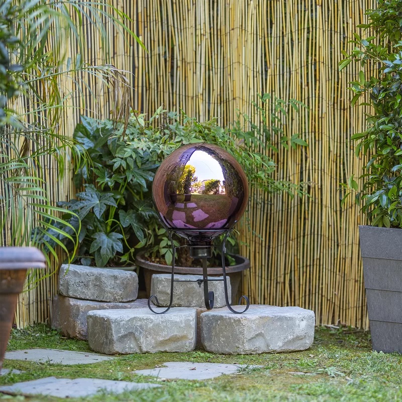 A reflective garden sphere on a metal stand, set against a bamboo background with surrounding greenery.