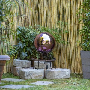 A reflective garden sphere on a metal stand, set against a bamboo background with surrounding greenery.