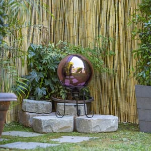 A reflective garden sphere on a metal stand, set against a bamboo background with surrounding greenery.