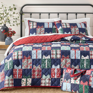 A patchwork quilt with matching pillow shams featuring a gift box design in red, blue, green, and plaid patterns is on a metal-framed bed. A tote bag with the same design is placed nearby, and vases with greenery and red berries sit on a side table.