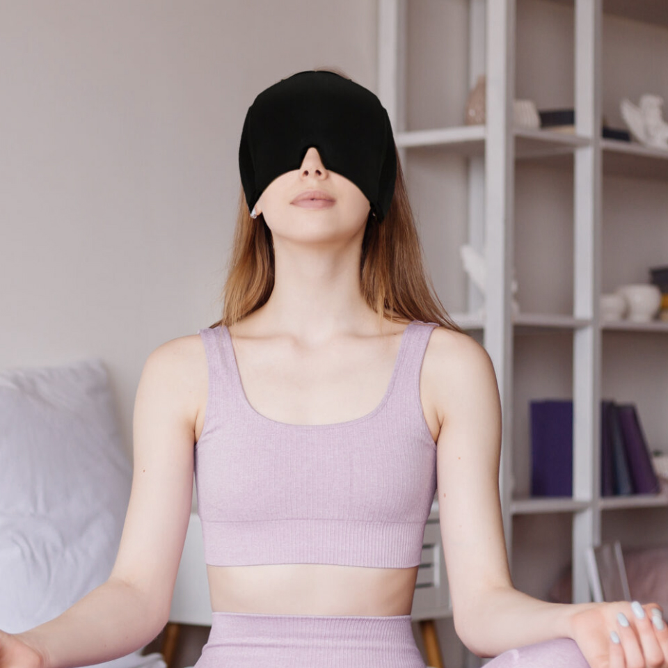 A woman wearing a black hot/cold head wrap relaxes with it covering her eyes. The wrap is suitable for soothing headaches or tension, allowing hands-free relief.