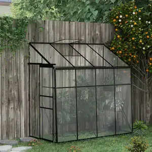 A small glass greenhouse with a black frame is positioned against a wooden fence, near a fruit-bearing tree and surrounded by grass.