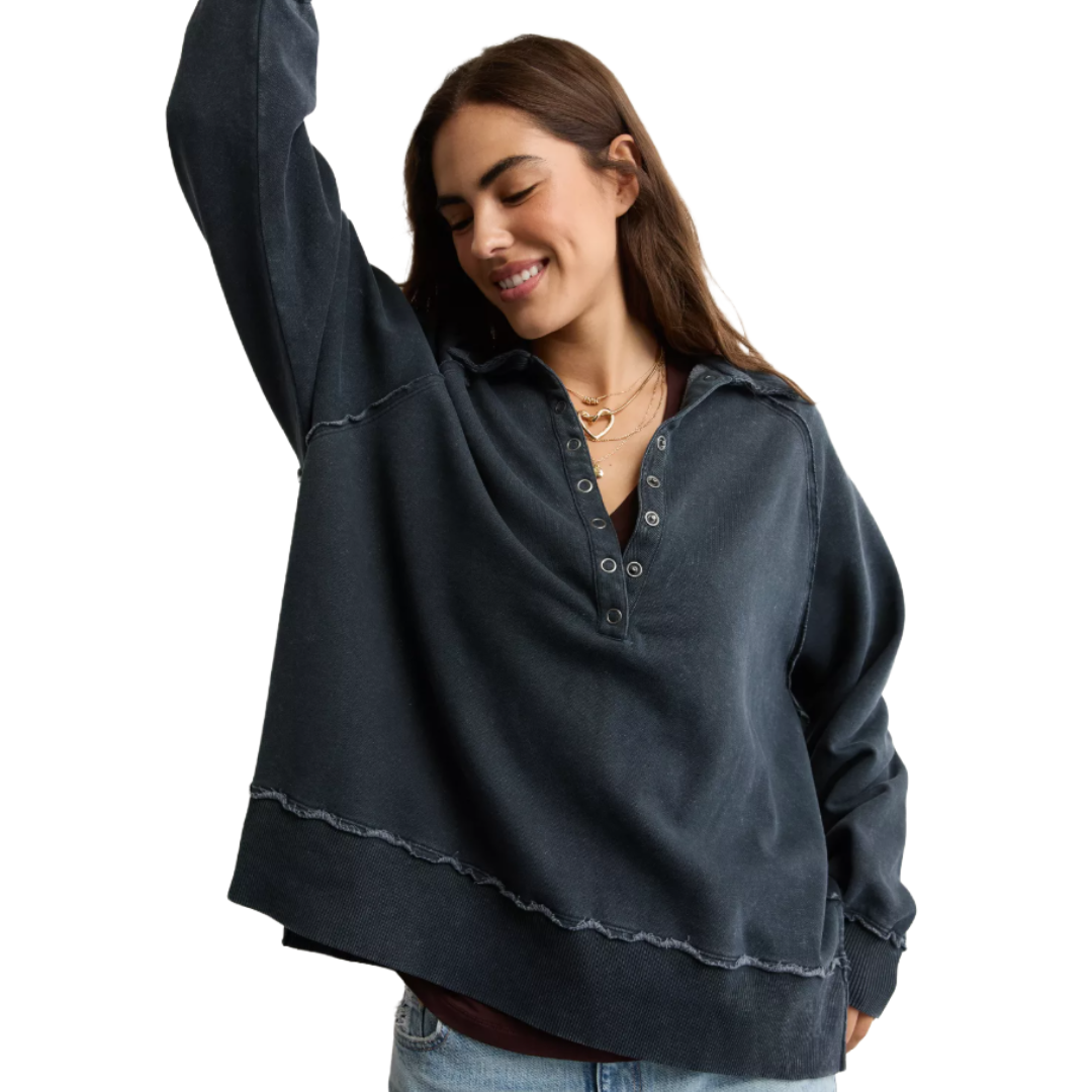 A woman wearing a dark, oversized henley-style pullover with visible seam details, paired with light blue jeans. She has layered necklaces, including one with a heart pendant.
