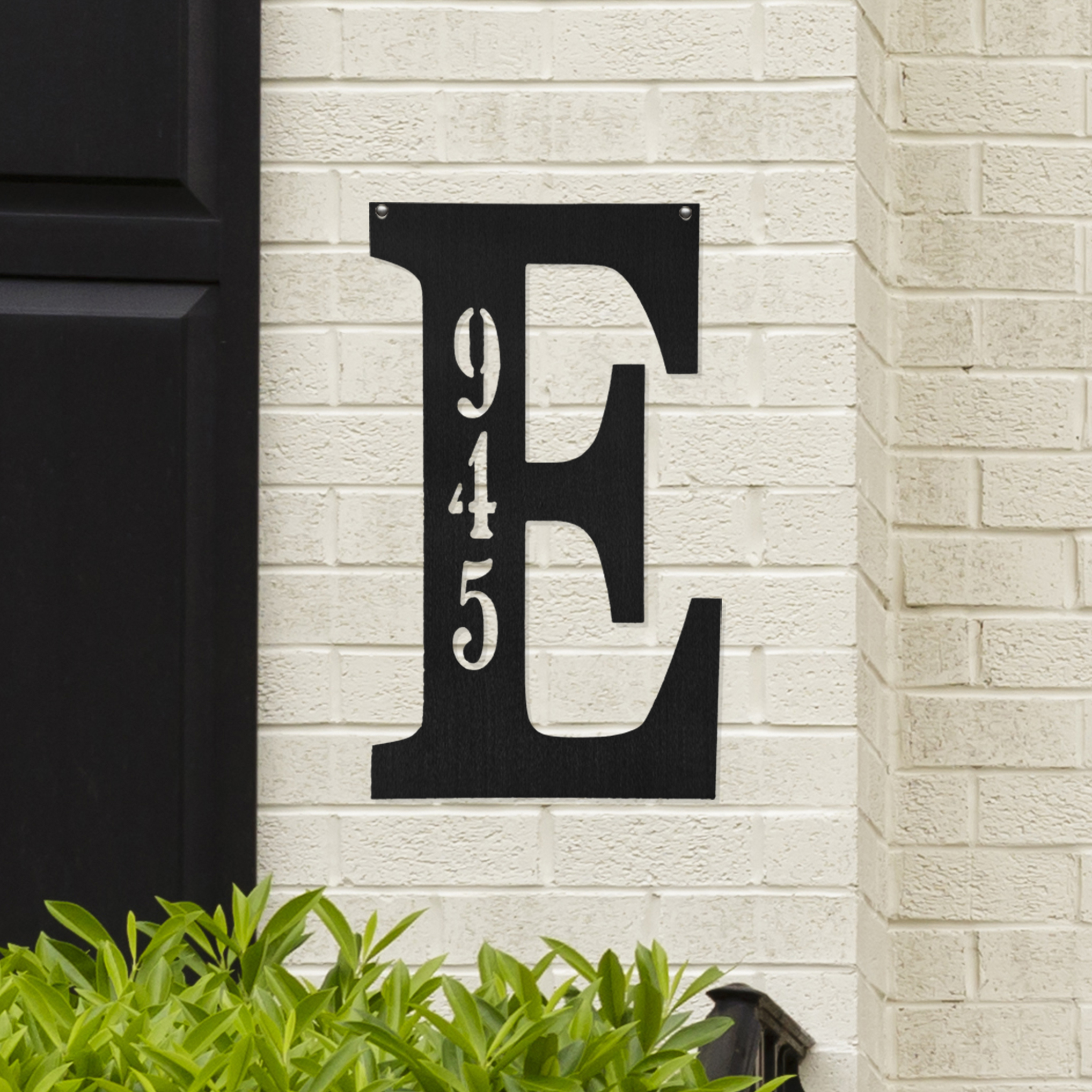 A large black letter \“E\“ sign with the numbers \“945\“ vertically inscribed is mounted on a light brick wall, with greenery visible at the bottom.