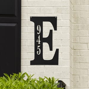 A large black letter \“E\“ sign with the numbers \“945\“ vertically inscribed is mounted on a light brick wall, with greenery visible at the bottom.