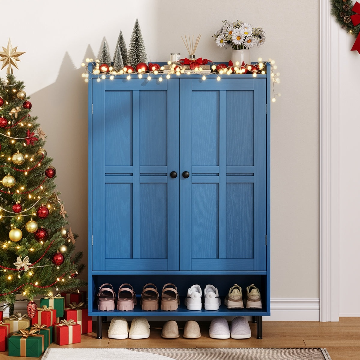 A blue shoe cabinet with decorative lights, mini trees, and flowers on top is next to a Christmas tree with ornaments and gifts. Shoes and slippers are neatly arranged below the cabinet.