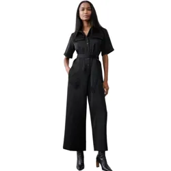 A woman is wearing a black short-sleeved jumpsuit with a button-up front and a belt at the waist. She pairs it with black ankle boots.