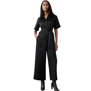 A woman is wearing a black short-sleeved jumpsuit with a button-up front and a belt at the waist. She pairs it with black ankle boots.