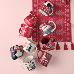 A collection of various Christmas-themed mugs with festive designs and text like \“Naughty,\“ \“Nice,\“ and \“Meowy Christmas.\“