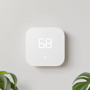 White square digital thermostat displaying a temperature of 68 degrees, mounted on a wall near a green plant leaf.