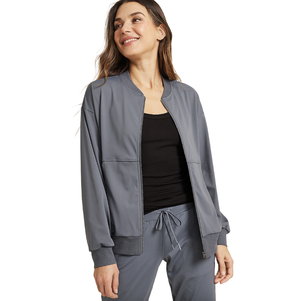 The Women's Traveler Jacket is a lightweight, long-sleeve zipper jacket in a soft gray color, featuring a ribbed collar and cuffs. It is paired with matching drawstring pants and worn over a black tank top.