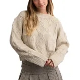 A woman is wearing a cream-colored, cable-knit sweater with ribbed cuffs and a pleated houndstooth skirt.