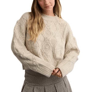 A woman is wearing a cream-colored, cable-knit sweater with ribbed cuffs and a pleated houndstooth skirt.