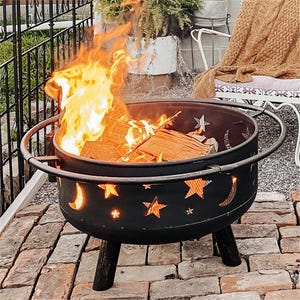 A fire pit with star and moon cutouts, emitting flames, is set on a paver patio with a chair and blanket nearby.