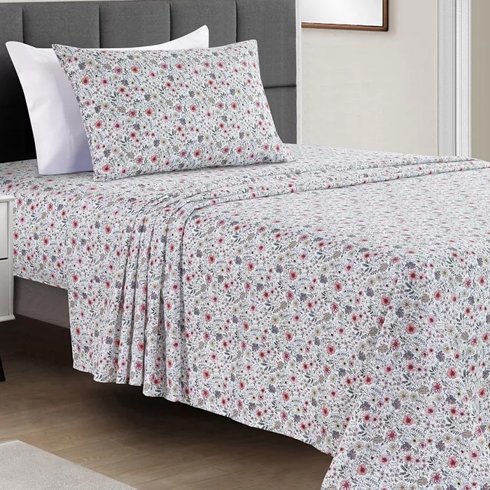 A bed set featuring floral-patterned sheets and pillowcases with pink, red, and green flowers on a white background, arranged on a neatly made bed with a gray upholstered headboard.